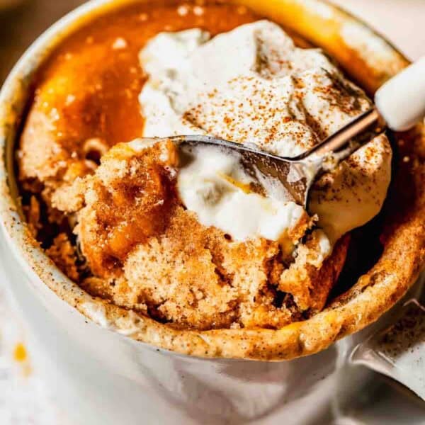 A close-up of a mug filled with cinnamon spiced mug cake, topped with a dollop of whipped cream and a sprinkle of cinnamon, with a spoon scooping out a portion.