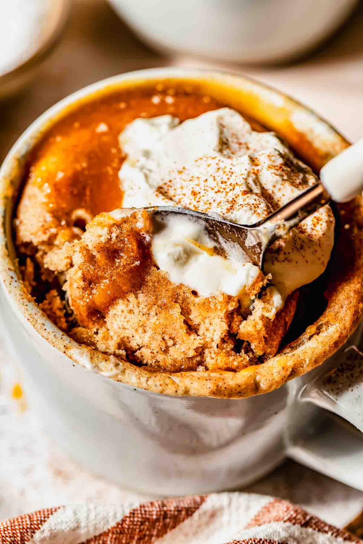 A close-up of a mug filled with cinnamon spiced mug cake, topped with a dollop of whipped cream and a sprinkle of cinnamon, with a spoon scooping out a portion.