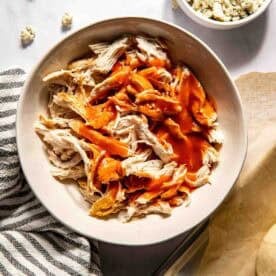 A bowl of shredded chicken topped with buffalo sauce, with a bowl of crumbled blue cheese and a striped towel nearby.