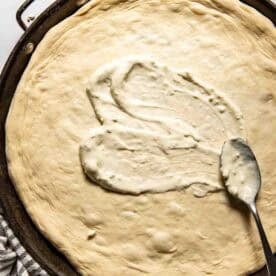 A large round pizza dough in a pan with blue cheese dressing being spread on it with a spoon.