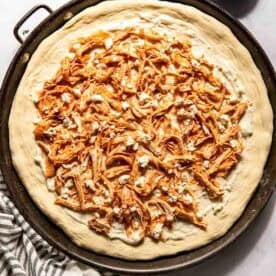 Unbaked pizza on a round tray topped with shredded buffalo chicken and crumbled cheese, with a bowl of cheese and a striped towel nearby.