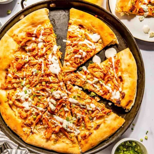 A baked pizza with shredded buffalo chicken, cheese, and white sauce on a round pan, surrounded by a drink, a sliced serving on a plate, green onions, sauce, and a striped towel.