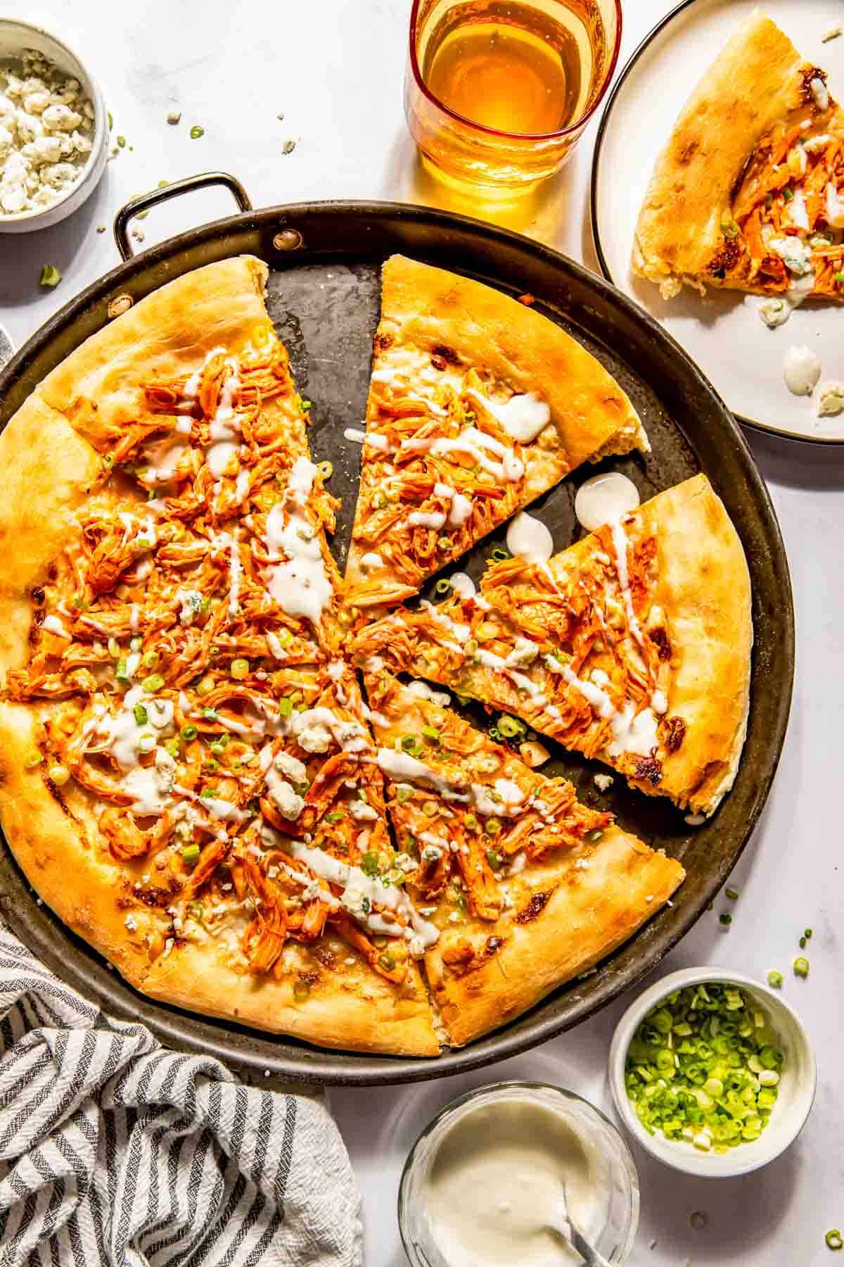 A baked pizza with shredded buffalo chicken, cheese, and white sauce on a round pan, surrounded by a drink, a sliced serving on a plate, green onions, sauce, and a striped towel.