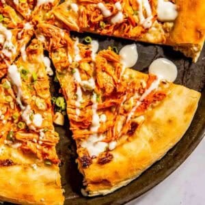 A pizza topped with shredded buffalo chicken, sliced green onions, and a drizzle of white sauce, cut into slices on a round pan.
