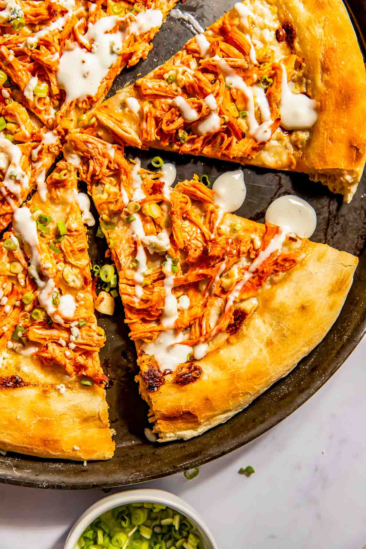 A pizza topped with shredded buffalo chicken, sliced green onions, and a drizzle of white sauce, cut into slices on a round pan.