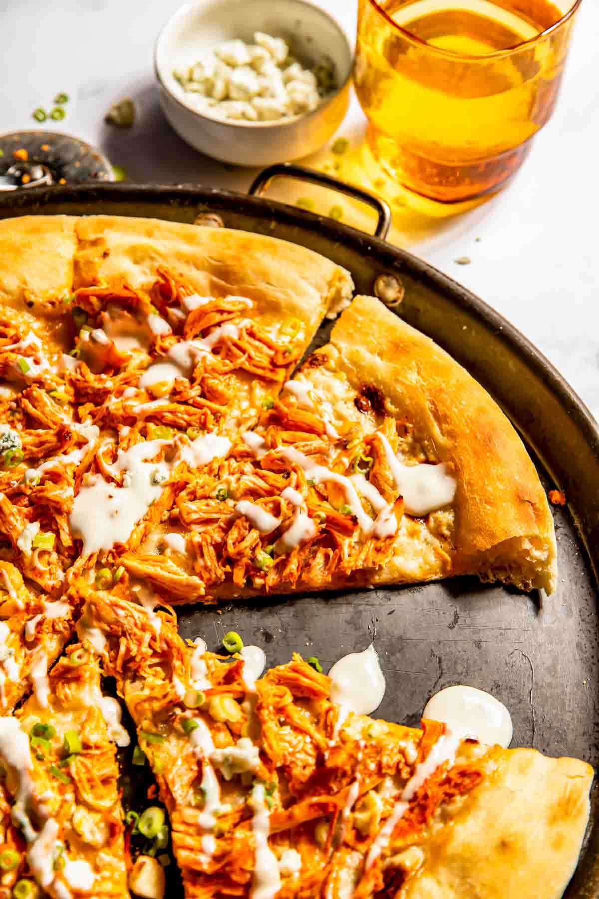 A baked pizza topped with shredded buffalo chicken, drizzled with white sauce, sliced on a round baking tray, with a glass of amber drink and a bowl of crumbled cheese in the background.