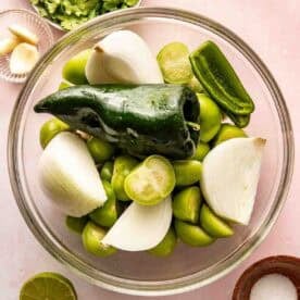 A glass bowl filled with tomatillos, halved onions, a poblano pepper, and a jalapeño. Surrounding the bowl are cilantro, garlic cloves, lime halves, and a small bowl of salt.