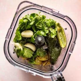 A blender filled with fresh cilantro, garlic cloves, jalapeño, and other green ingredients, viewed from above.