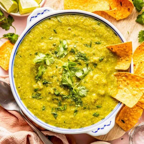 A bowl of green salsa garnished with cilantro, surrounded by tortilla chips, lime wedges, and a small bowl of chopped cilantro on a wooden board.