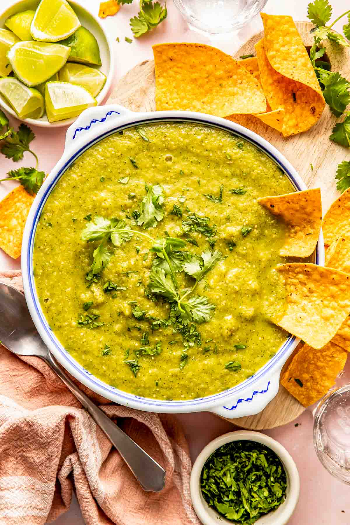 A bowl of green salsa garnished with cilantro, surrounded by tortilla chips, lime wedges, and a small bowl of chopped cilantro on a wooden board.