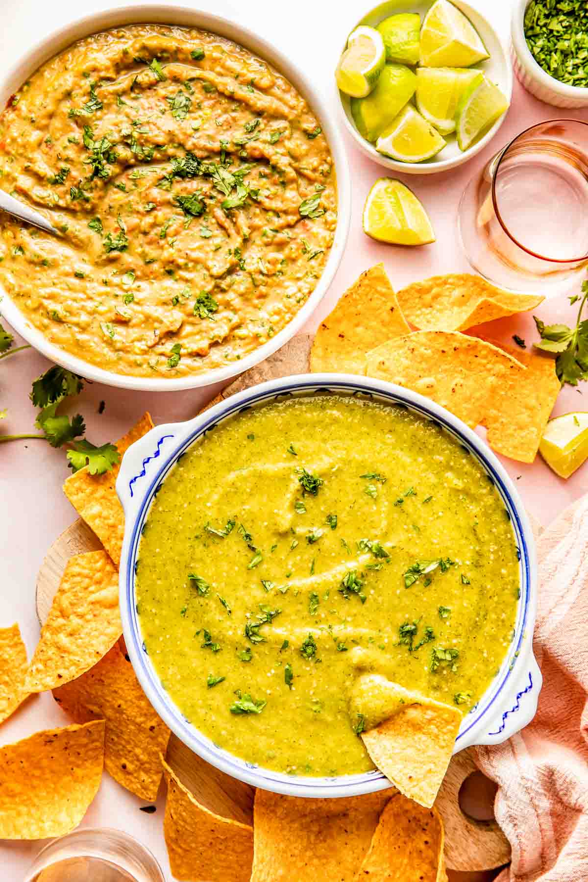 Two bowls of green and yellow dips garnished with herbs, surrounded by tortilla chips, lime wedges, and fresh cilantro on a white surface.