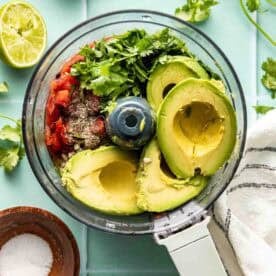 Food processor filled with avocado, tomatoes, cilantro, spices, and lime halves nearby, on a light blue surface with a striped towel and a small bowl of salt.