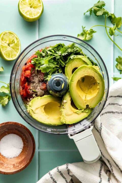 Food processor filled with avocado, tomatoes, cilantro, spices, and lime halves nearby, on a light blue surface with a striped towel and a small bowl of salt.