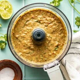 A food processor containing blended salsa sits on a light green surface with halved limes, cilantro, a bowl of salt, and a striped towel nearby.
