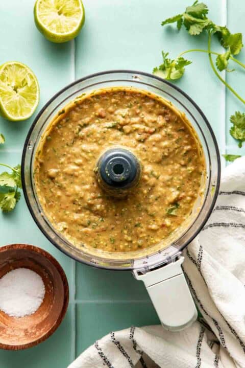 A food processor containing blended salsa sits on a light green surface with halved limes, cilantro, a bowl of salt, and a striped towel nearby.