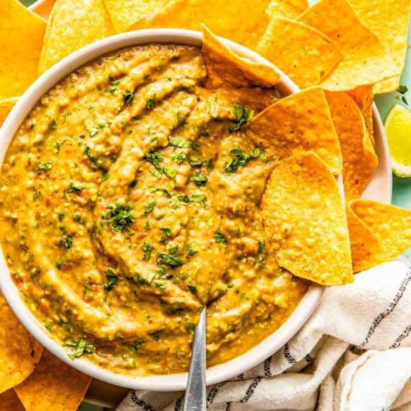 A bowl of creamy salsa garnished with chopped herbs, surrounded by tortilla chips, with a spoon in the bowl and a striped cloth nearby.