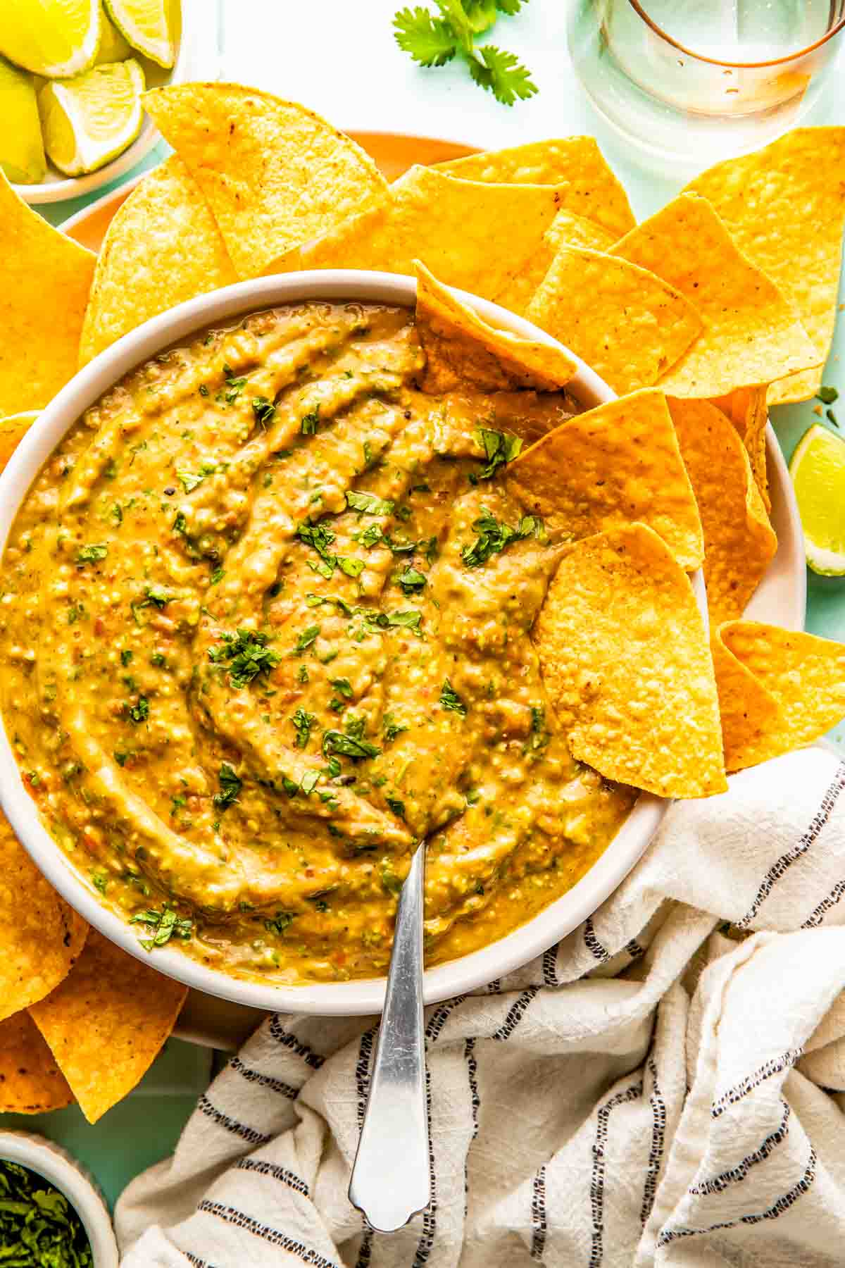 A bowl of creamy salsa garnished with chopped herbs, surrounded by tortilla chips, with a spoon in the bowl and a striped cloth nearby.