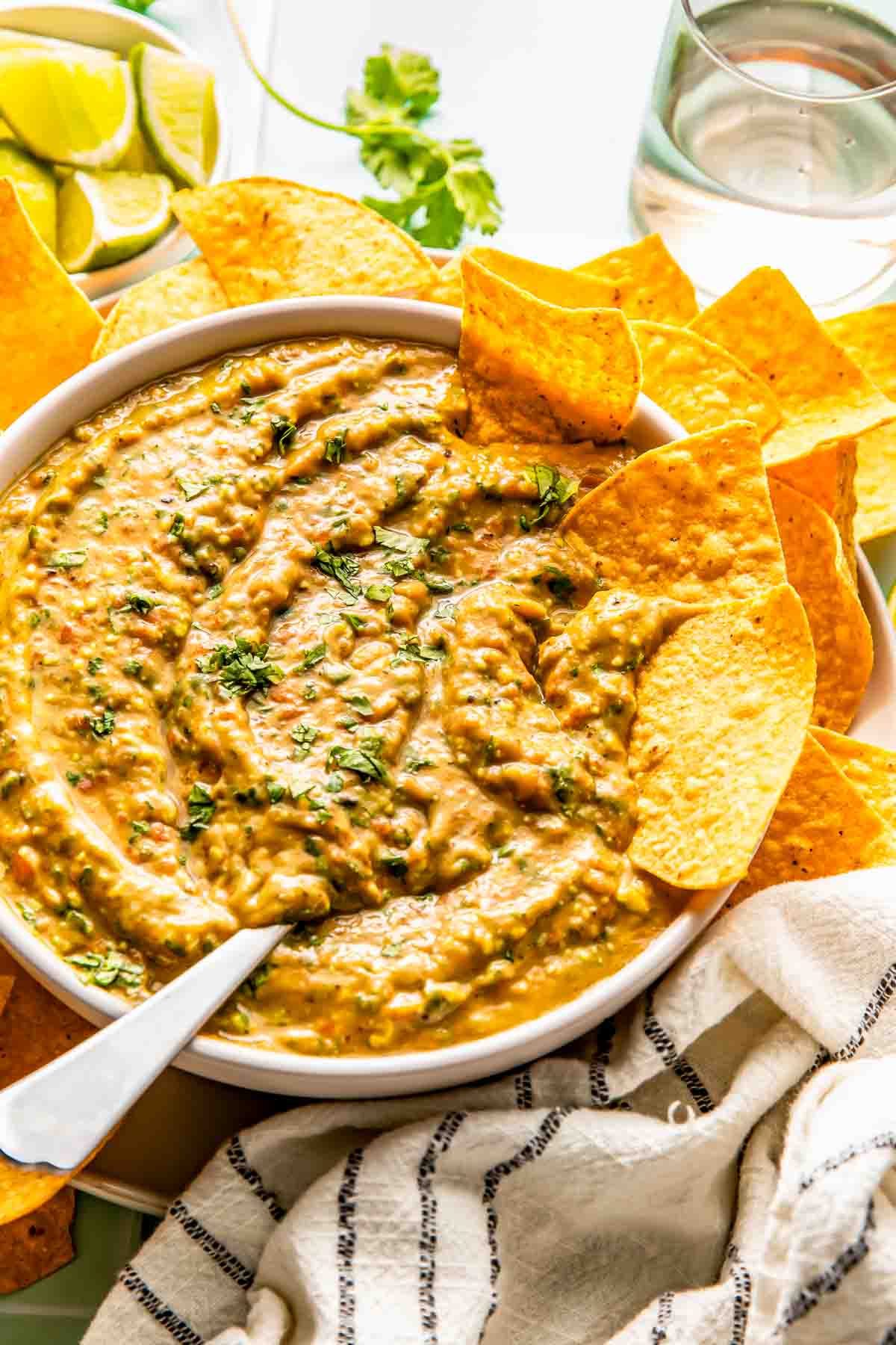 A bowl of creamy salsa garnished with herbs, surrounded by tortilla chips, with lime wedges and a glass of water in the background.