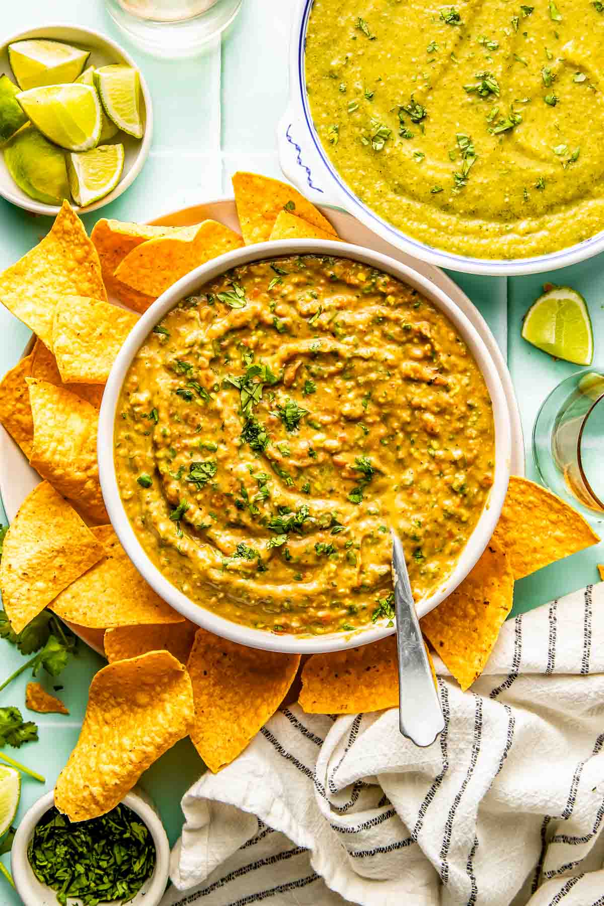 A bowl of creamy salsa garnished with herbs, surrounded by tortilla chips, with lime wedges and another bowl of green dip nearby.