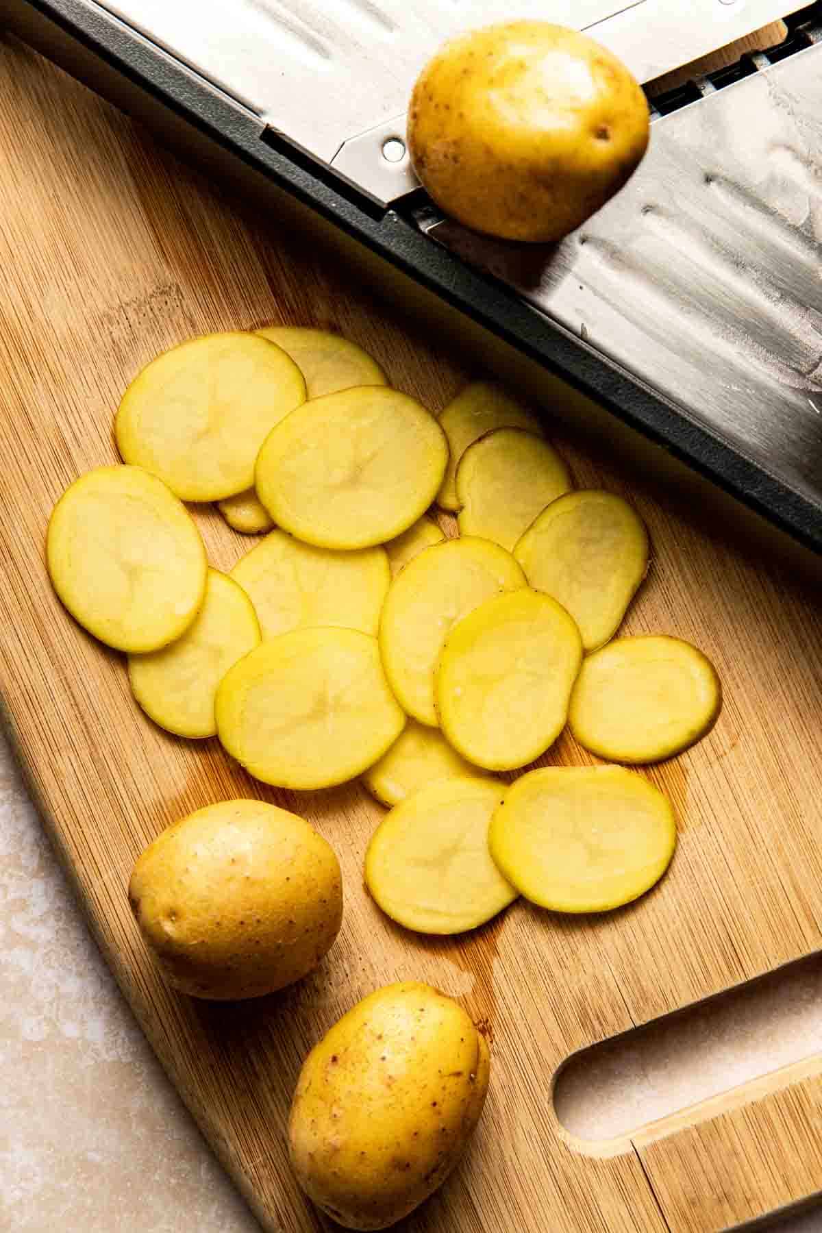 Sliced yellow potatoes on a wooden cutting board next to a mandoline slicer and whole potatoes.