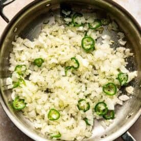 Chopped onions and sliced jalapeños sautéing in a stainless steel pan.