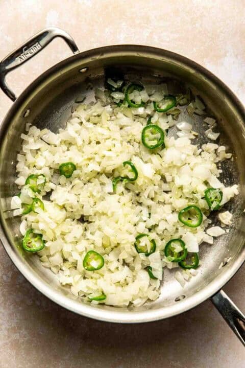 Chopped onions and sliced jalapeños sautéing in a stainless steel pan.
