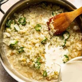 A saucepan with chopped onions and jalapeños sautéing, a wooden spoon, and cream being poured in from a measuring cup.
