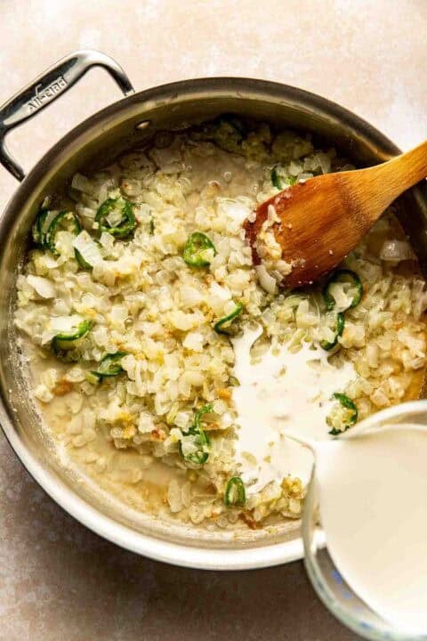 A saucepan with chopped onions and jalapeños sautéing, a wooden spoon, and cream being poured in from a measuring cup.