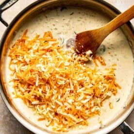 A metal pan filled with creamy sauce and shredded cheese being stirred with a wooden spoon.