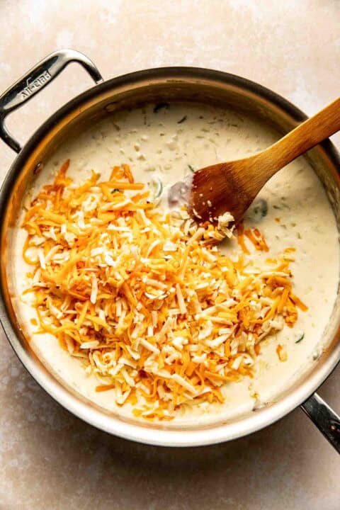 A metal pan filled with creamy sauce and shredded cheese being stirred with a wooden spoon.