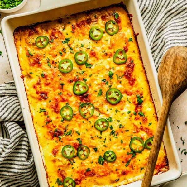 A baked casserole topped with melted cheese and sliced jalapeños in a rectangular white dish, with a wooden spoon and bowls of herbs and jalapeños nearby.