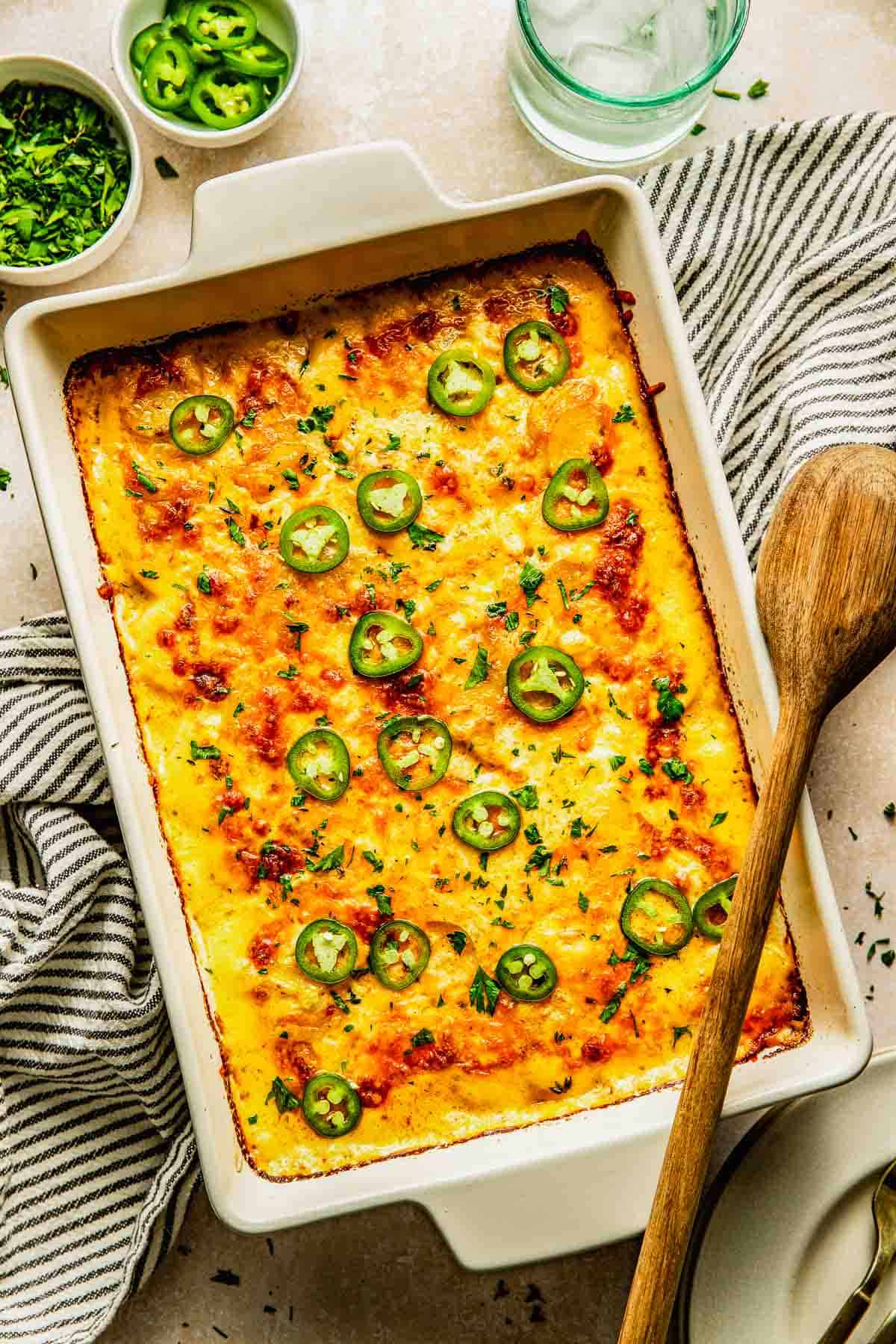 A baked casserole topped with melted cheese and sliced jalapeños in a rectangular white dish, with a wooden spoon and bowls of herbs and jalapeños nearby.