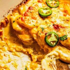Close-up of a cheesy baked casserole topped with sliced jalapenos, with a wooden spoon scooping out a portion, showing creamy and melted cheese layers.