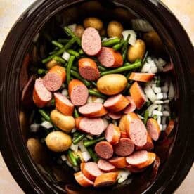 Sliced sausage, baby potatoes, green beans, and chopped onions in a slow cooker, ready to be cooked.