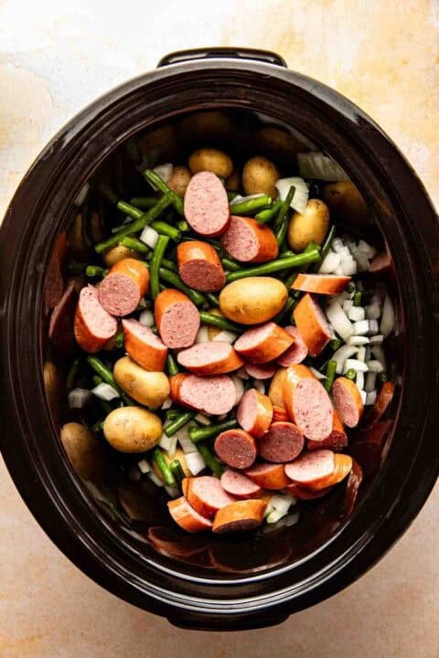 Sliced sausage, baby potatoes, green beans, and chopped onions in a slow cooker, ready to be cooked.