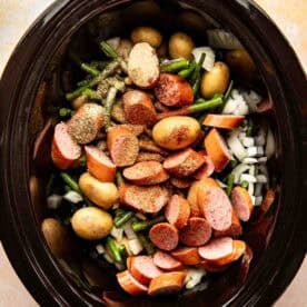 Sliced sausage, baby potatoes, green beans, and chopped onions with seasoning inside a slow cooker, ready to be cooked.