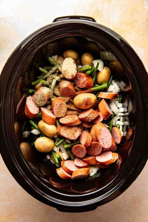 Sliced sausage, baby potatoes, green beans, and chopped onions with seasoning inside a slow cooker, ready to be cooked.