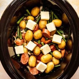 A slow cooker filled with whole baby potatoes, sliced sausage, green beans, onion pieces, and cubes of butter, ready to be cooked.