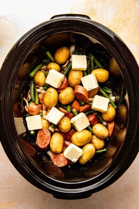 A slow cooker filled with whole baby potatoes, sliced sausage, green beans, onion pieces, and cubes of butter, ready to be cooked.