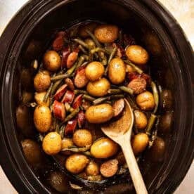 A slow cooker filled with small potatoes, green beans, sausage slices, and a wooden spoon.