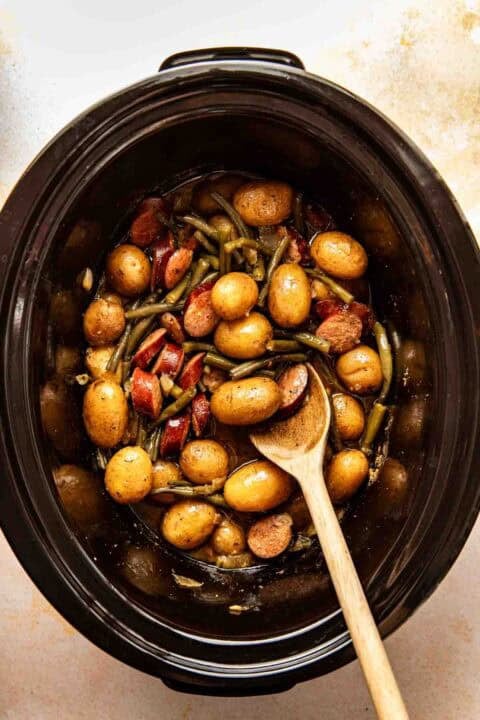 A slow cooker filled with small potatoes, green beans, sausage slices, and a wooden spoon.