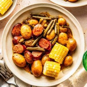 A bowl containing baby potatoes, green beans, sausage slices, and corn on the cob, served on a beige surface with utensils and other plates nearby.