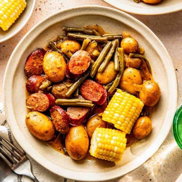 A bowl containing baby potatoes, green beans, sausage slices, and corn on the cob, served on a beige surface with utensils and other plates nearby.
