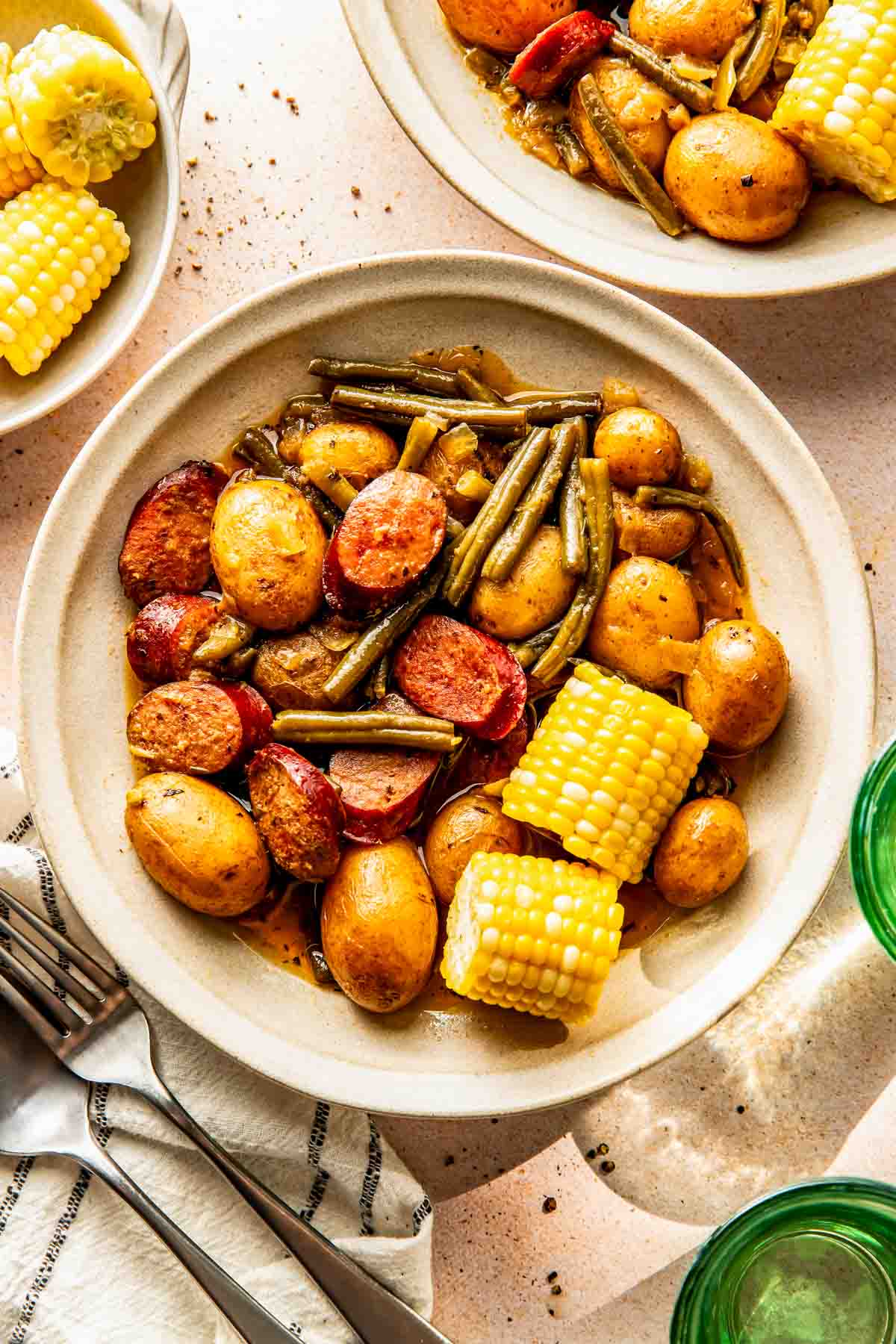 A bowl containing baby potatoes, green beans, sausage slices, and corn on the cob, served on a beige surface with utensils and other plates nearby.