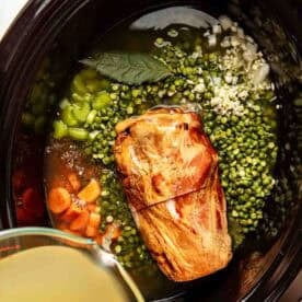 A slow cooker filled with a ham shank, split green peas, chopped carrots, celery, minced garlic, a bay leaf, and broth being poured in.