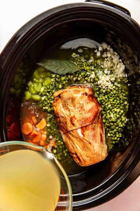 A slow cooker filled with a ham shank, split green peas, chopped carrots, celery, minced garlic, a bay leaf, and broth being poured in.