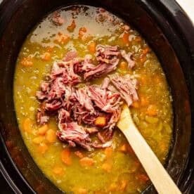 A slow cooker filled with split pea soup, visible carrots, and shredded ham being stirred with a wooden spoon.