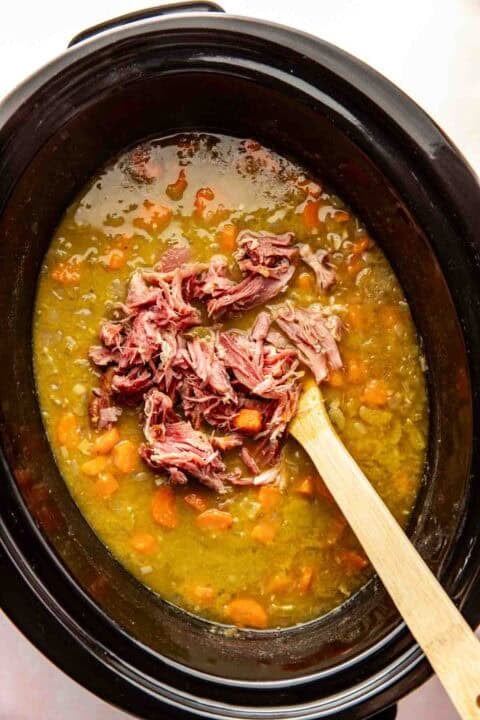 A slow cooker filled with split pea soup, visible carrots, and shredded ham being stirred with a wooden spoon.