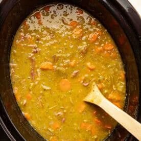 A slow cooker filled with split pea soup containing chopped carrots and ham, with a wooden spoon resting inside.