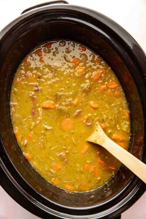 A slow cooker filled with split pea soup containing chopped carrots and ham, with a wooden spoon resting inside.